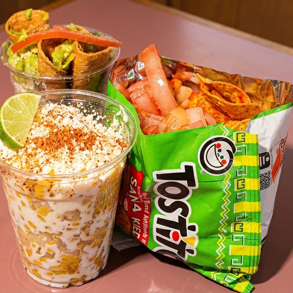 Elote, a Tostitos bag with toppings, and a cup of taquitos on a pink table.