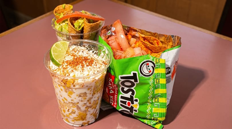 Elote, a Tostitos bag with toppings, and a cup of taquitos on a pink table.