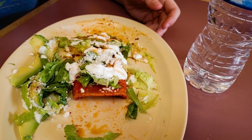 Partially eaten enchilada with lettuce, avocado, sour cream, and cheese on a yellow plate.