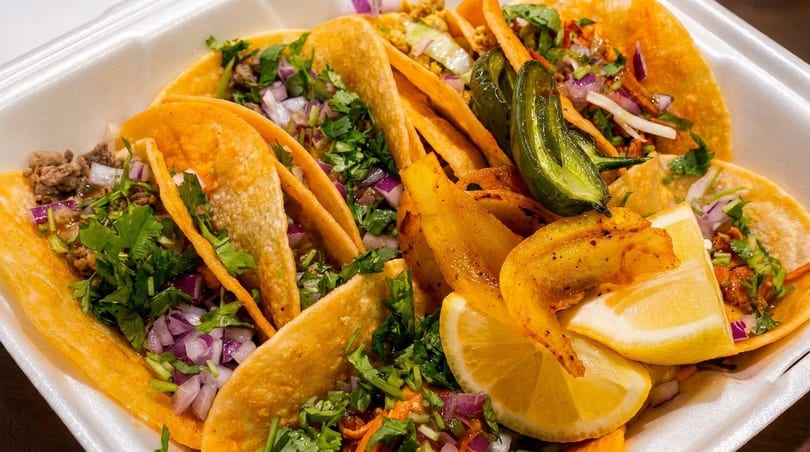 A white takeout container filled with several tacos, garnished with cilantro, red onion, lemon, and a jalapeño.